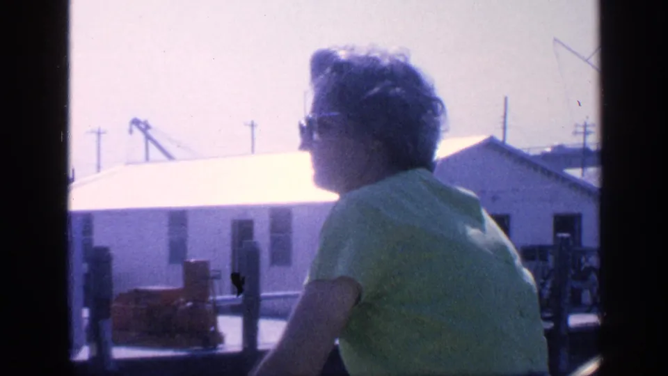 In supplied 1968 Florida 8mm footage, a woman in sunglasses and a green shirt rides past low waterfront buildings.