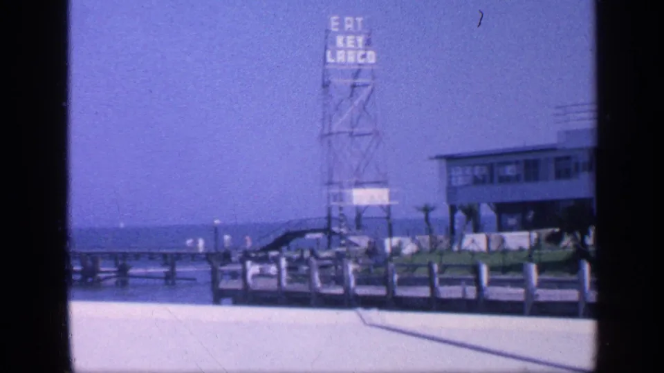 1968 8mm Florida beach view showing a pier area, Eat Key Largo sign, stilted waterfront building, railings, and ocean beyond.