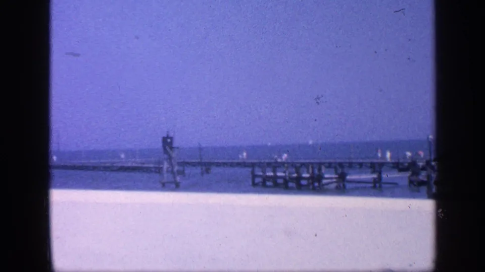 1968 8mm Florida beach view with long pier, ocean horizon, pale sand or walkway, railings, and a small tower structure.
