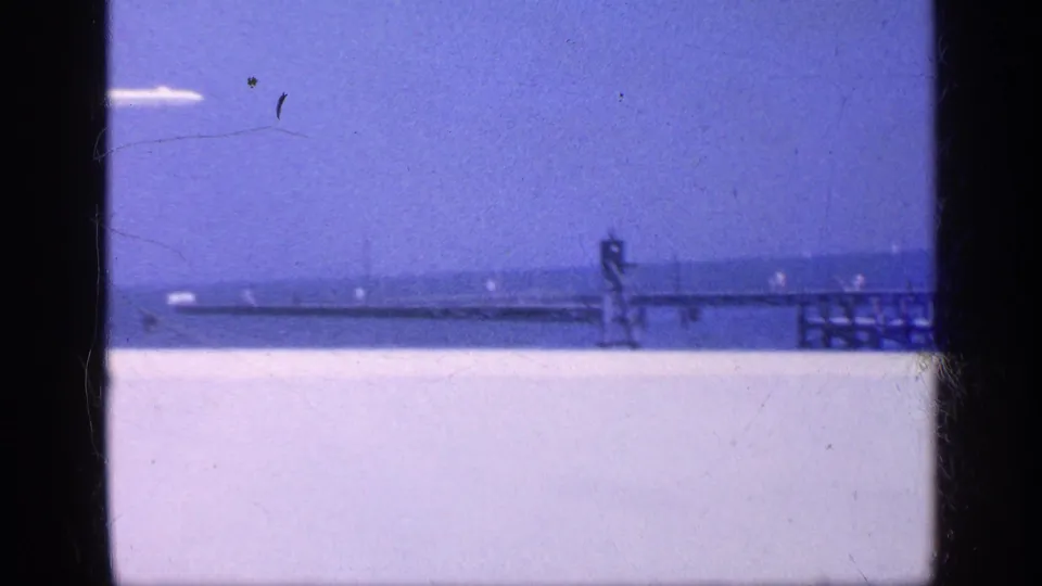 1968 Florida coastal frame showing the pier, ocean horizon, bright beach foreground, and a small airplane streaking across the sky.