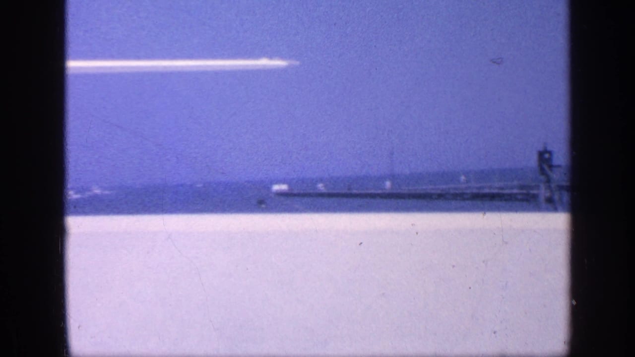 Open water with a distant pier or breakwater and a white aircraft crossing the sky.