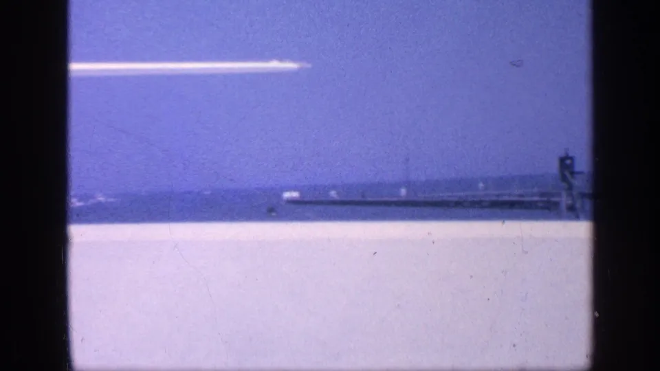 1968 Florida 8mm frame showing open water, a distant pier or breakwater, and a white aircraft against a blue sky.