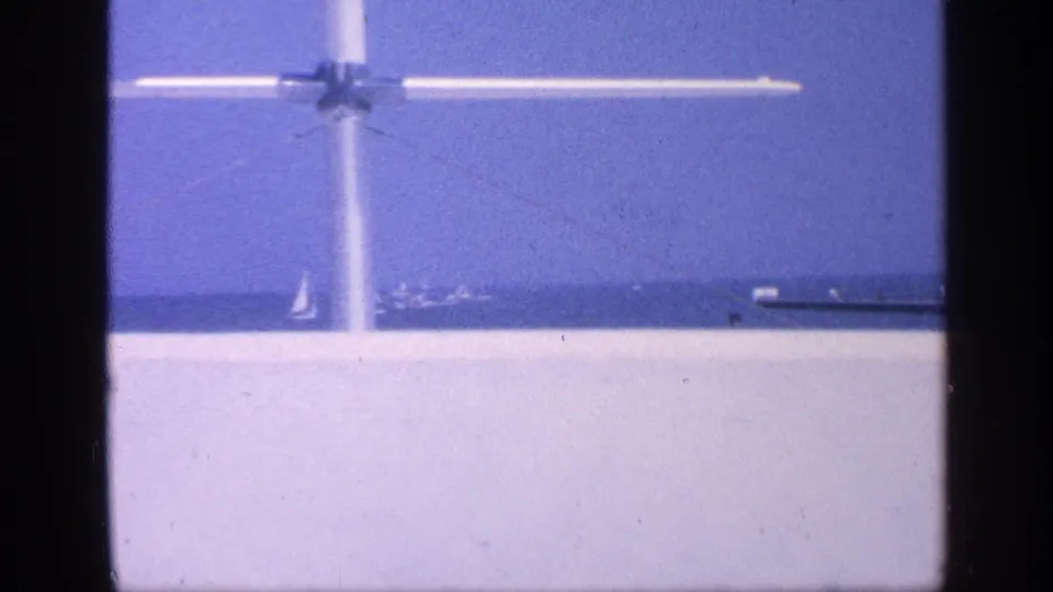 1968 Florida home-movie frame with a white aircraft overhead, open water, and distant shoreline or pier structures.