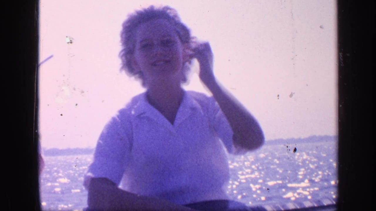 Woman in a white collared shirt sits by bright water, brushing back windblown hair.