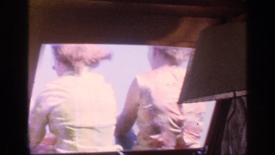 A home-movie view shows two adults from behind, seated closely within a dark-framed travel setting under strong daylight.