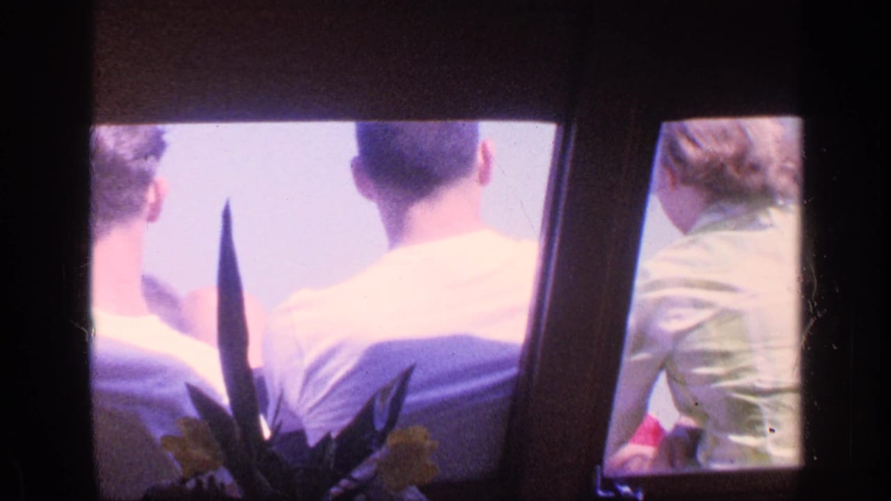 Two seated passengers in white shirts are viewed from behind through a dark boat interior frame.