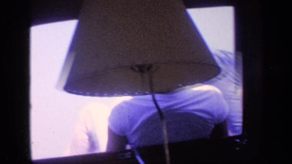 1968 Florida home-movie frame with a lampshade in the foreground and a male figure in a white T-shirt seen from behind at the windows.
