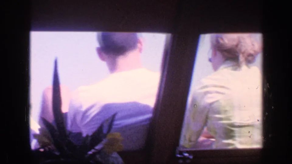 1968 Florida 8mm frame of seated passengers viewed from behind inside likely water transport, with bright exterior light and a plant silhouette.