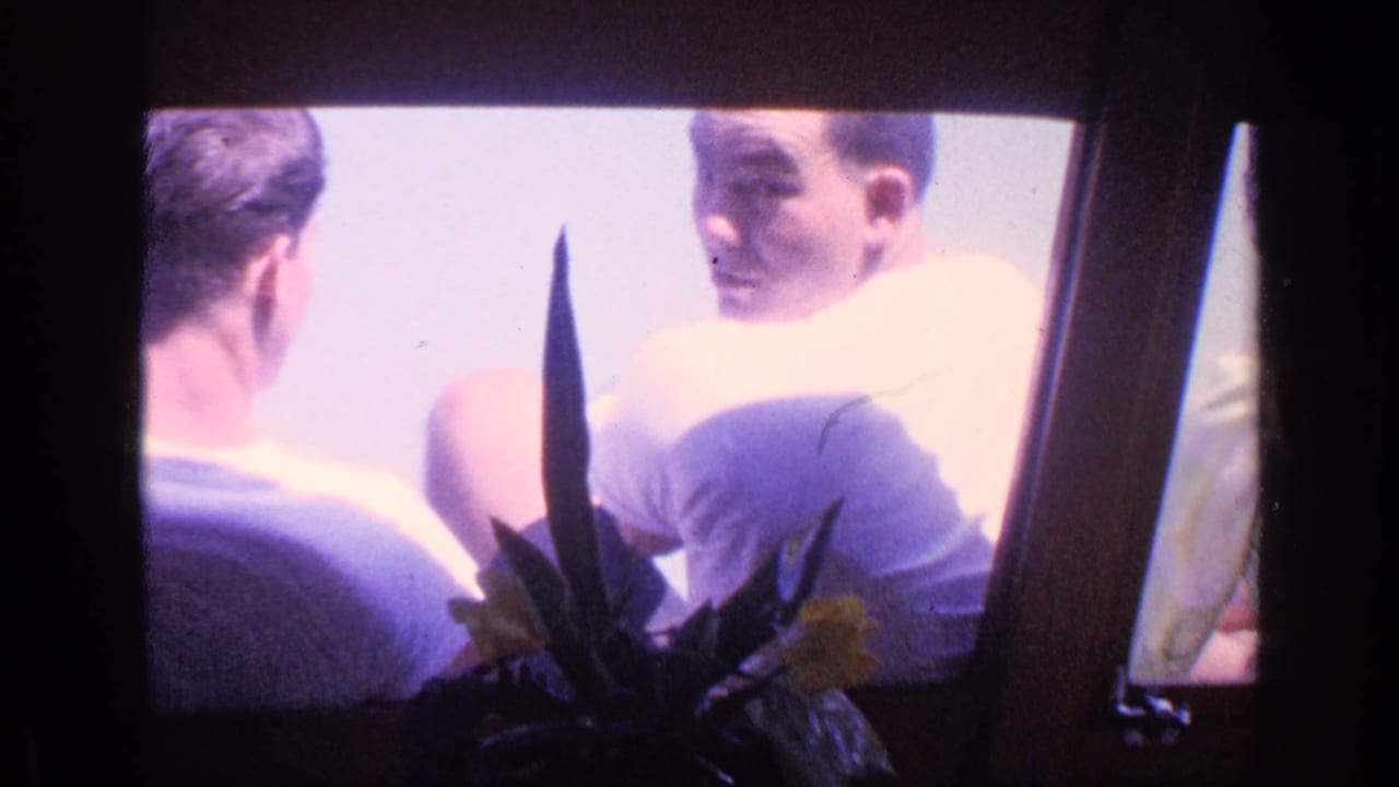 One seated man turns back and smiles while another man faces him near a bright window.