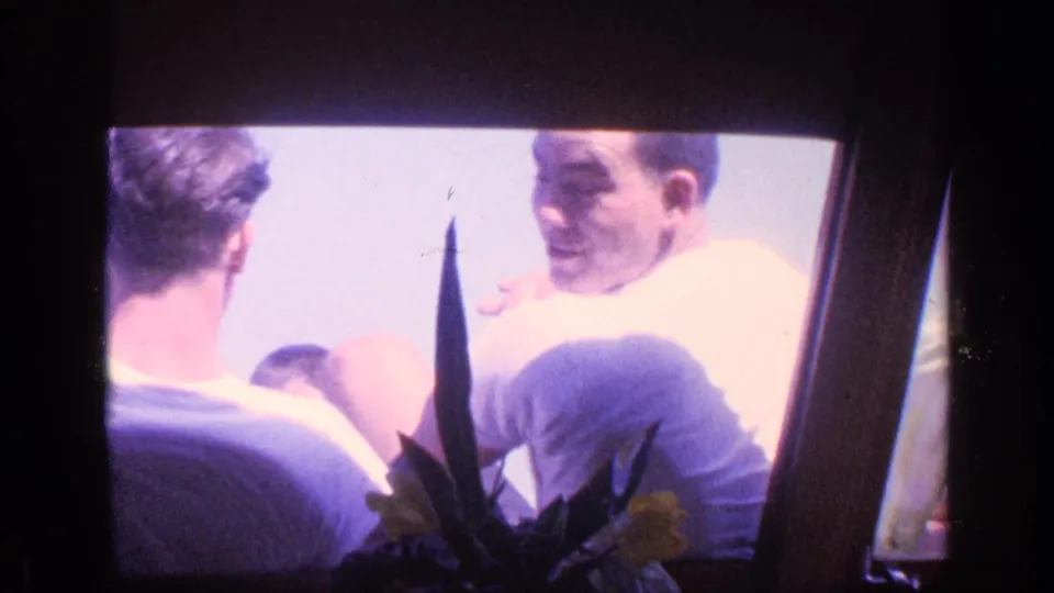 1968 Florida 8mm frame showing two seated young men near a bright window, one smiling back through soft exposure.