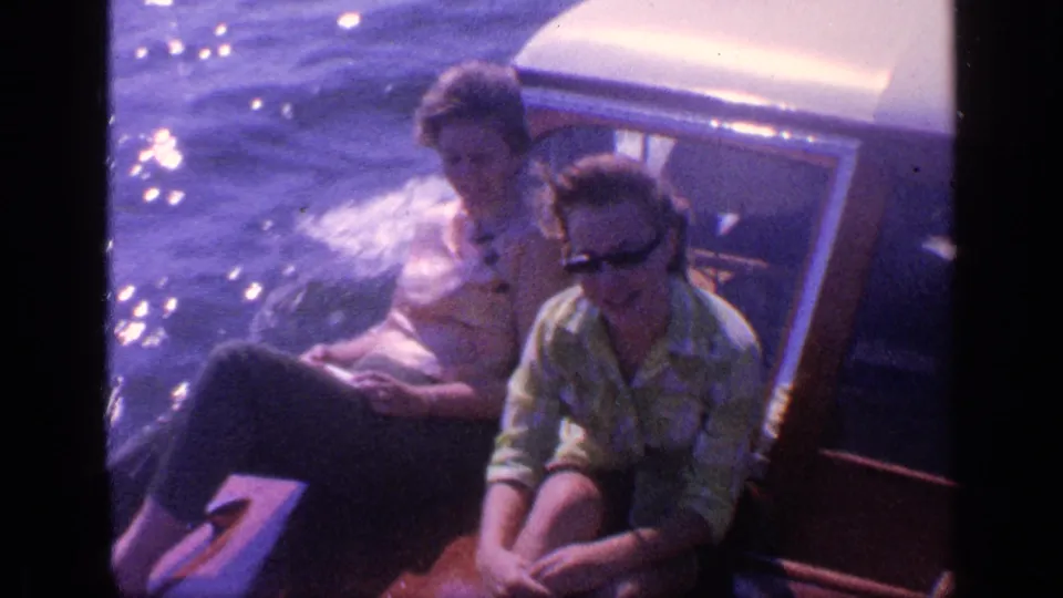 1968 Florida home-movie frame of two adults seated on a motorboat, with sunglasses, casual clothes, and sparkling water nearby.