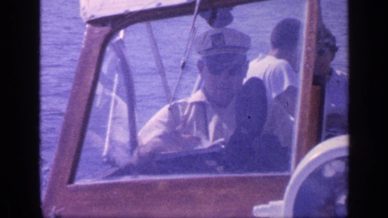 Man in captain hat steers a small wooden boat with passengers behind the windshield.