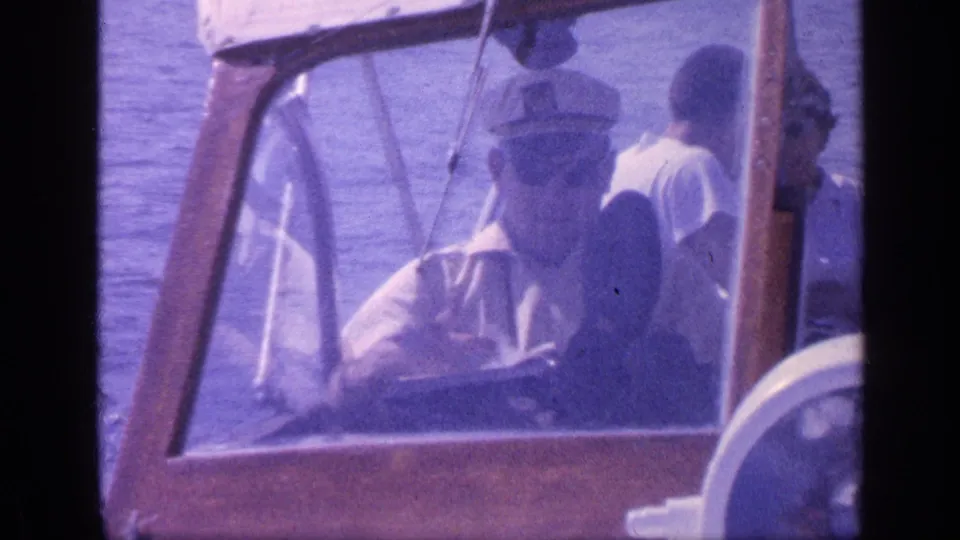 1968 Florida home-movie frame showing a man in a captain hat at the wheel of a small wooden boat with passengers nearby.
