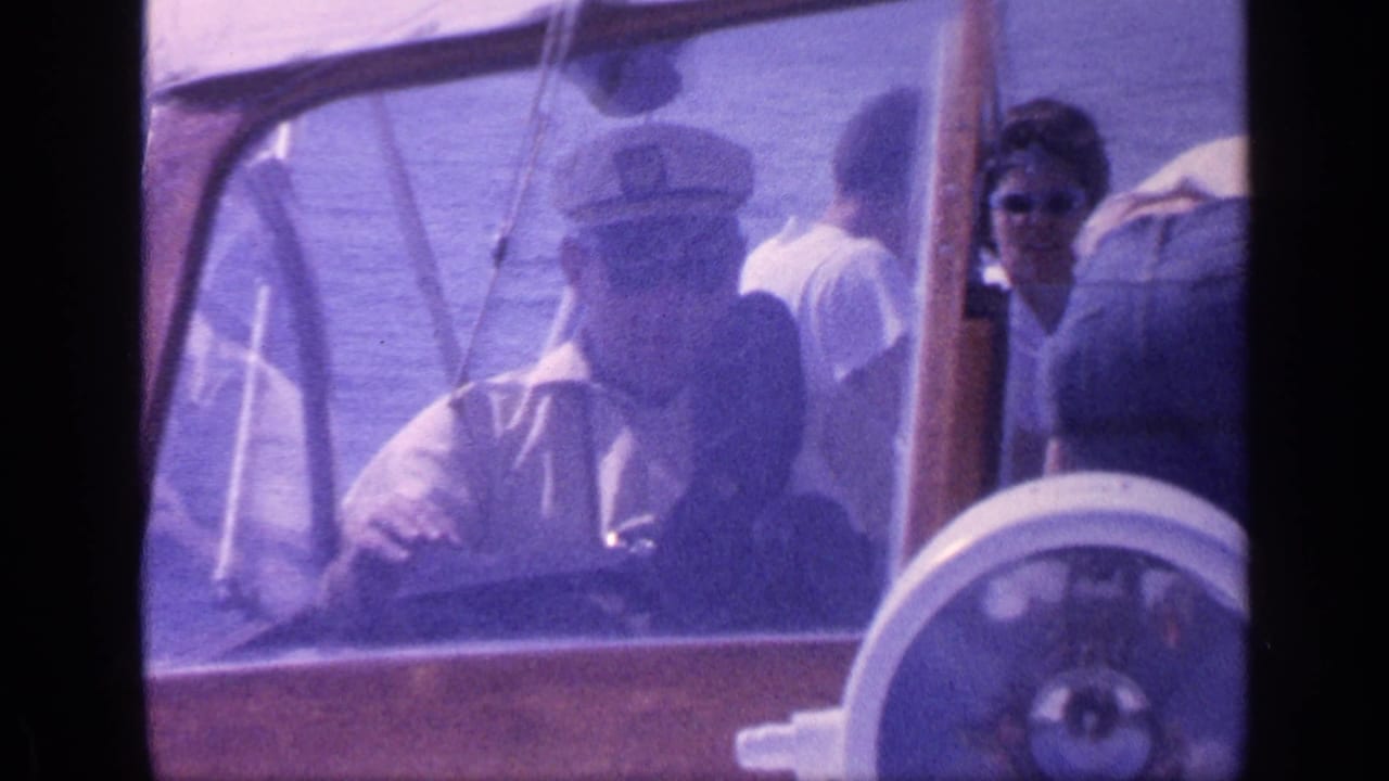 Driver in captain hat sits at the wheel while passengers stand and sit inside the boat.