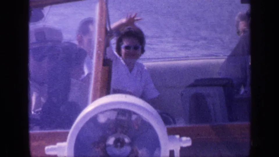 1968 Florida home-movie frame showing a woman in sunglasses waving from a small wooden boat with passengers around her.
