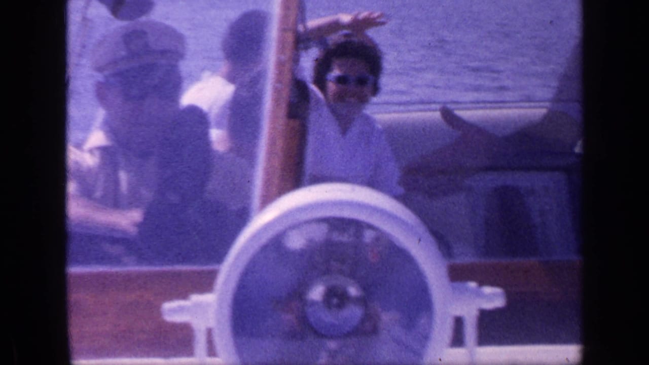 Passengers aboard a small motorboat, with a woman in sunglasses waving near a large white steering wheel.