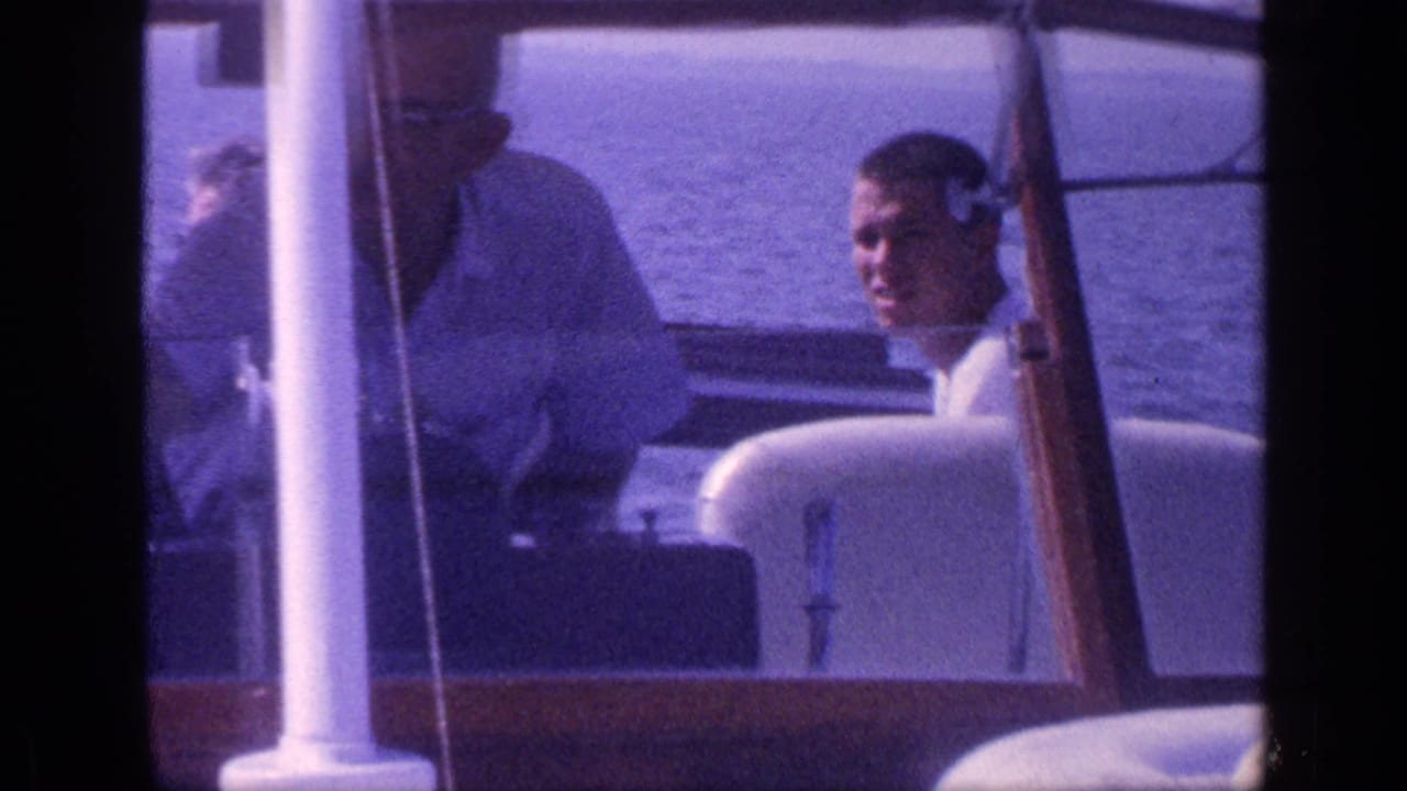 A man in a captain-style hat stands near the helm while another passenger sits on the motorboat.