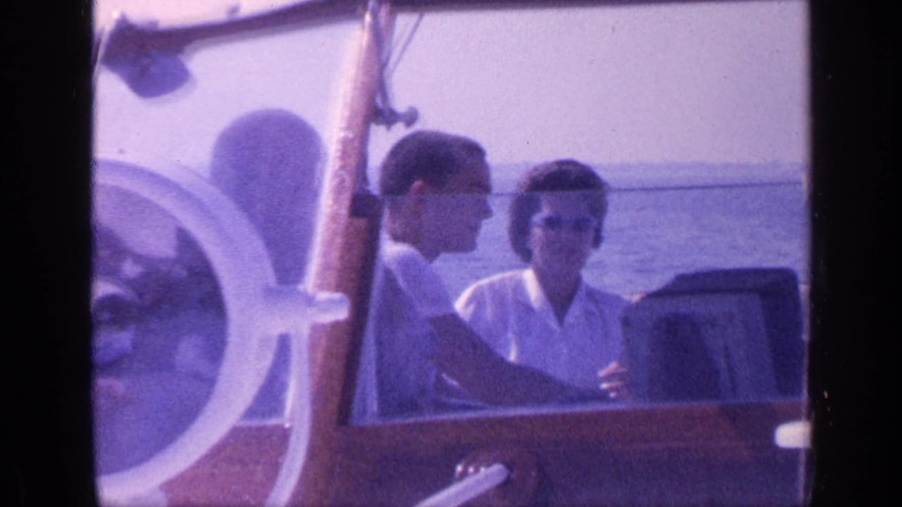 Two adults sit in a wooden boat near a white steering wheel on open water.