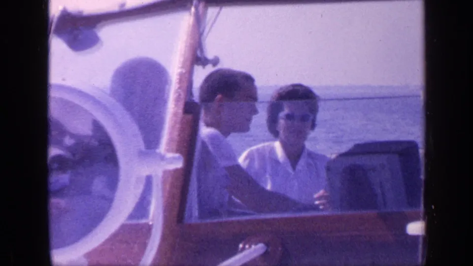 A man and woman sit together in a wooden boat in Florida, with sunglasses, a white steering wheel, and calm water visible.