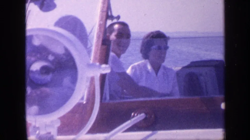 1968 Florida home-movie frame showing a couple seated in a wooden boat, partly framed by the white steering wheel and sunlit water.