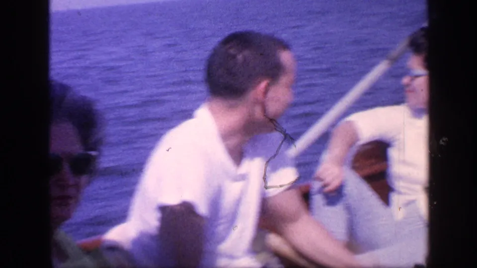 1968 Florida home-movie frame with a man in a white shirt seated near another passenger, framed closely against the blue ocean.