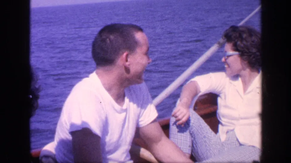 1968 Florida home-movie frame showing a man and woman facing each other on a boat, with a diagonal rail and ocean behind them.