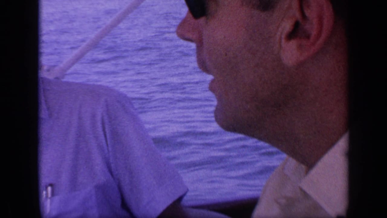 Close profile of a man in sunglasses on a boat with water and another shirted figure behind him.