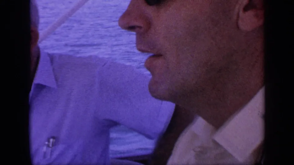 1968 Florida home-movie frame showing a close profile of a man in sunglasses on a boat, with a light-shirted passenger and water behind him.