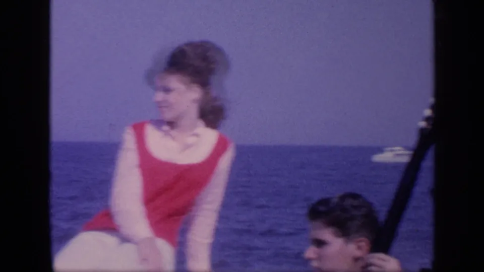 1968 Florida home-movie frame showing an adult woman in a red swimsuit seated on a boat, with a man nearby, open sea, and a dark guitar-like shape at the edge.