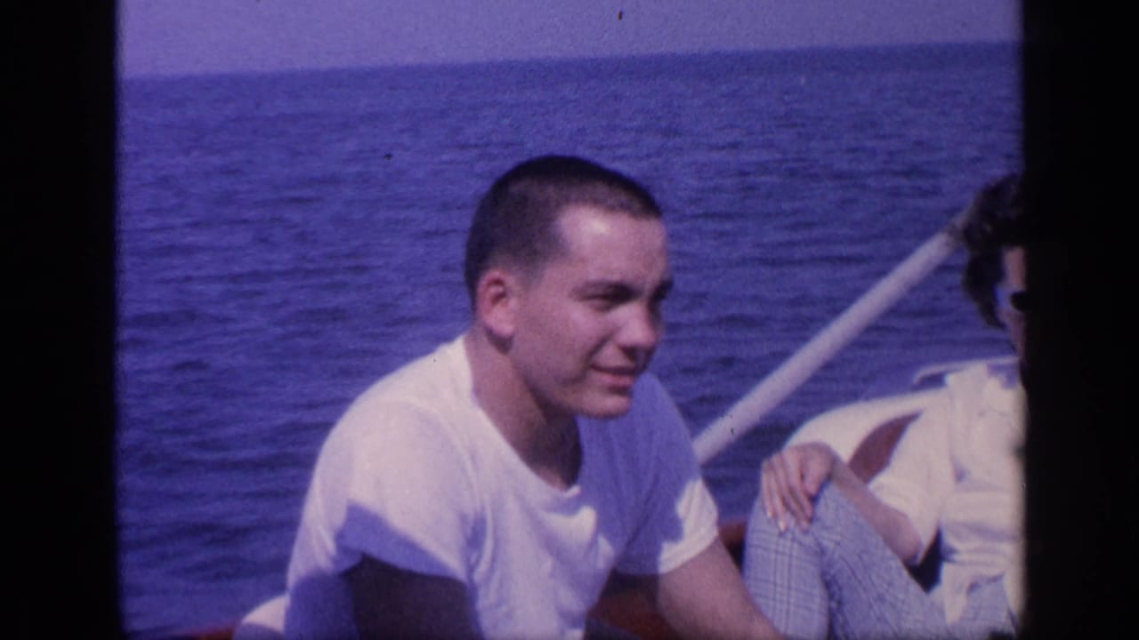 Young man in a white T-shirt seated on a boat with ocean behind him.