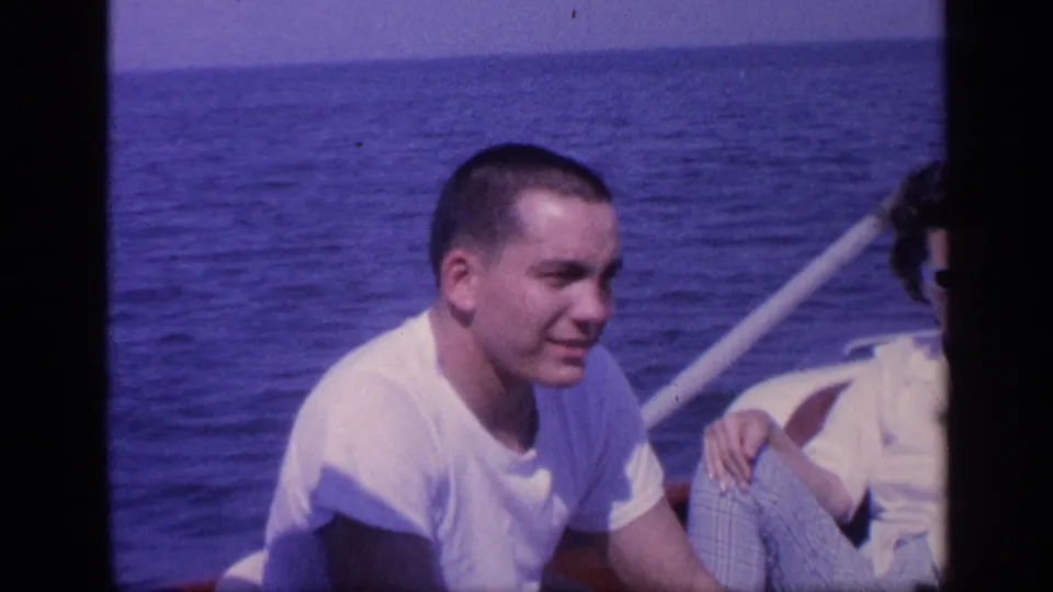 A young man in a white T-shirt sits on a boat off Florida, with blue ocean behind him and a companion partly visible nearby.