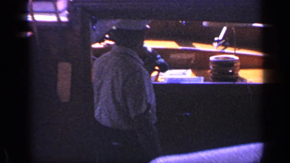 A person seen from behind stands or sits in a dim cabin-like interior, with a lit counter area and small objects ahead.