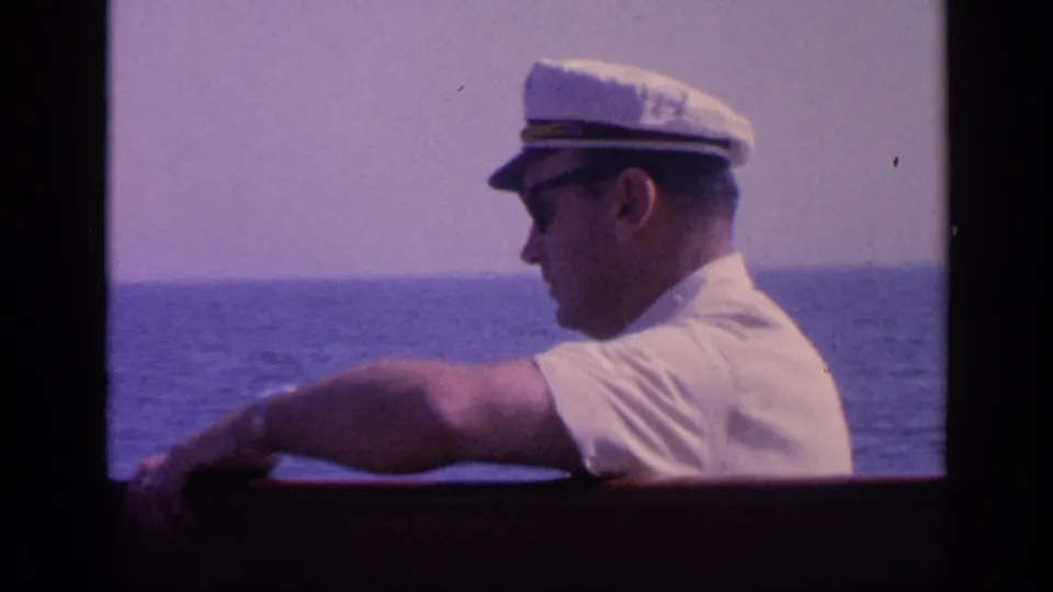 Side view of the same adult man in sunglasses and a white cap at the boat rail, with open water behind him in faded 8mm color.