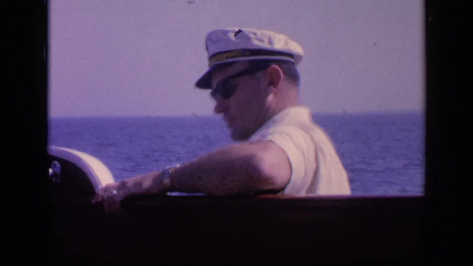 Adult man in a white captain's hat and sunglasses rests his arm on the boat rail and looks toward the sea in 1968 Florida home-movie footage.