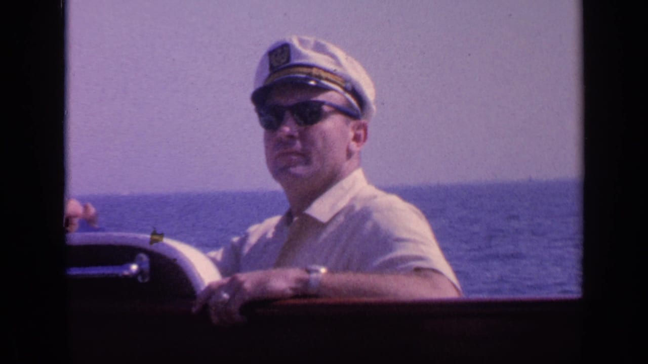 In 1968 Florida home-movie footage, a man in a white captain-style hat and sunglasses holds the boat wheel with open water behind him.