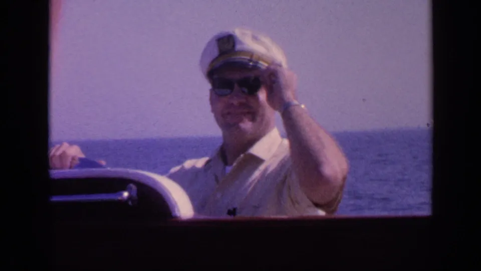 The man smiles and raises a hand toward the camera while seated by the boat controls in the Florida boating home movie.