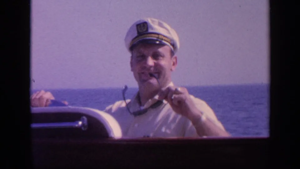 A man in a white captain-style hat holds his sunglasses near his face, smiling beside the boat controls with open water in the background.