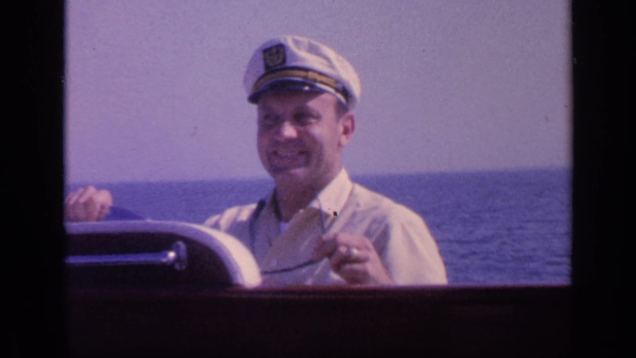 Smiling man in a white captain's hat steering a boat on open water.