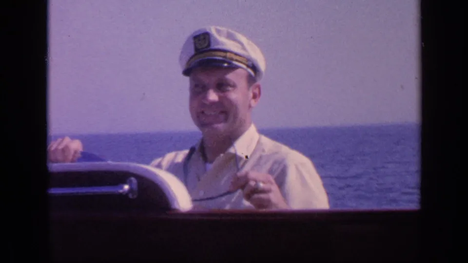 1968 Florida home-movie frame showing a smiling man in a white captain's hat at the helm of a boat on sunny open water.
