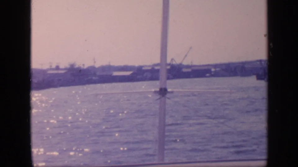 1968 Florida home-movie frame looking from a boat across sunlit water toward a low port shoreline, cranes, and bridge supports.