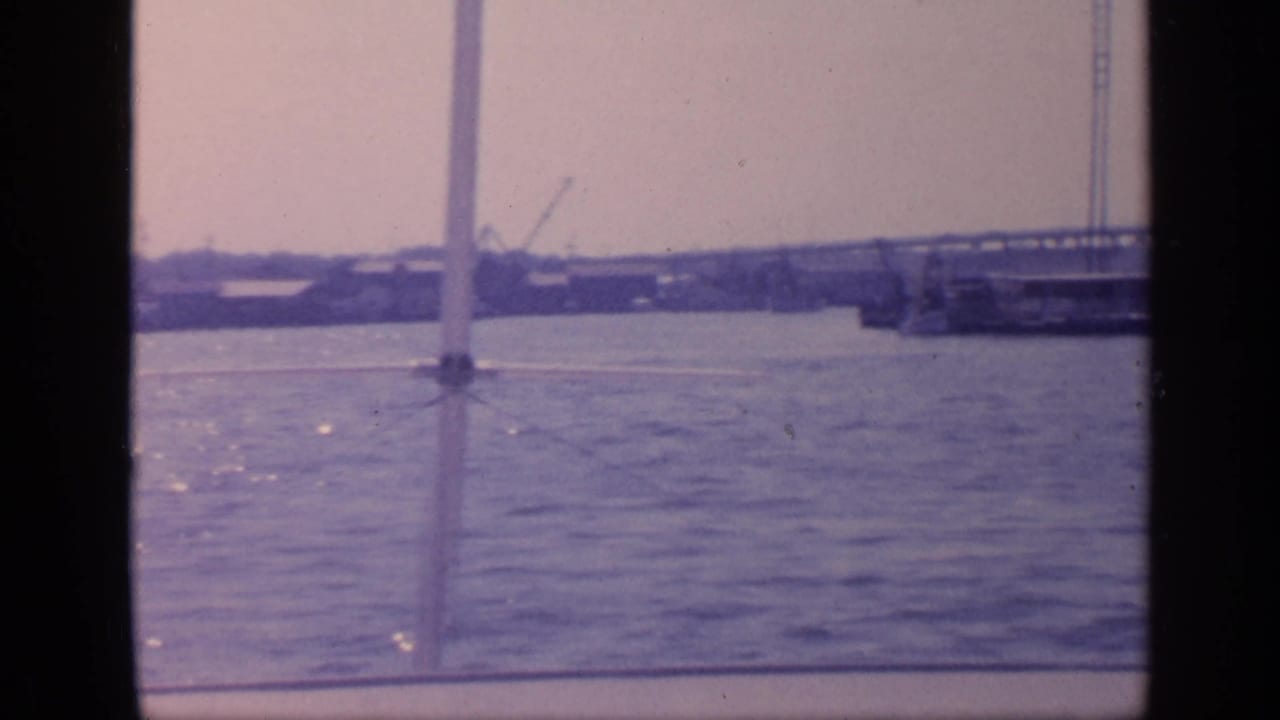 1968 Florida boat view with dark vertical framing, open water, a bridge span, and distant industrial shoreline structures.
