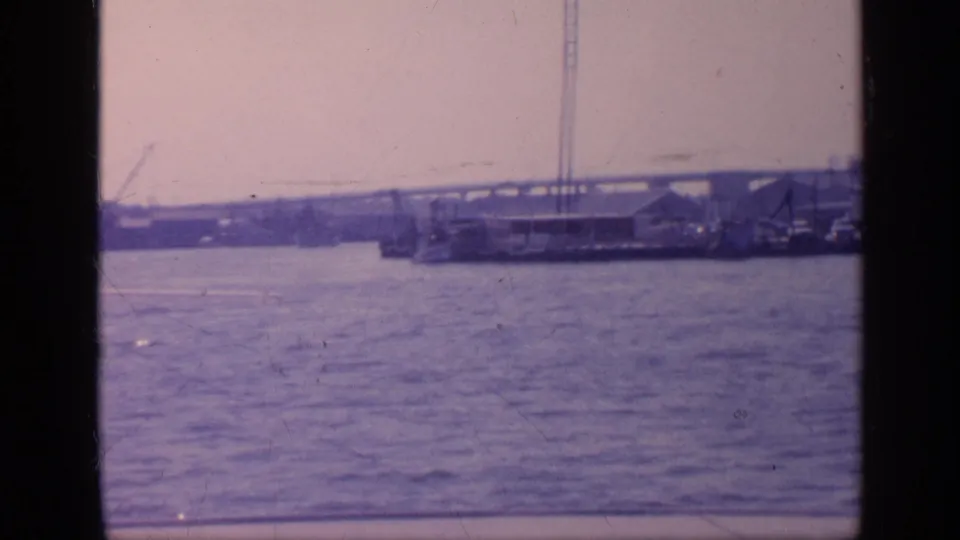 1968 Florida home-movie frame showing rippled water with a distant port edge, cranes, low structures, and part of a bridge support.