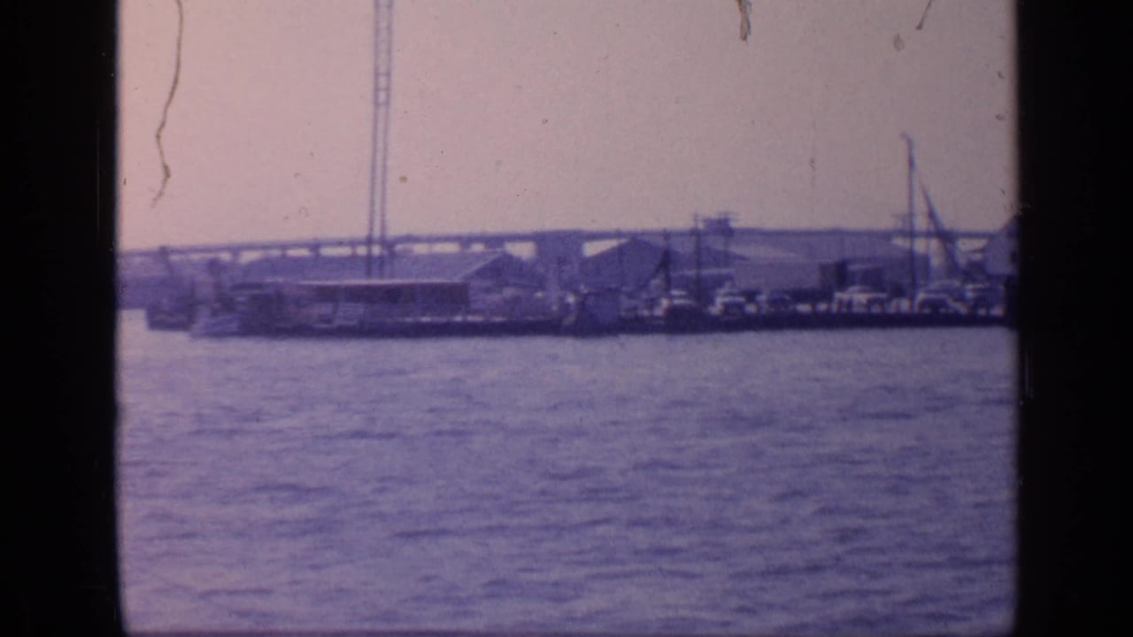 Wide harbor view with water, docked boats, industrial sheds, cranes, and a long bridge in the background.