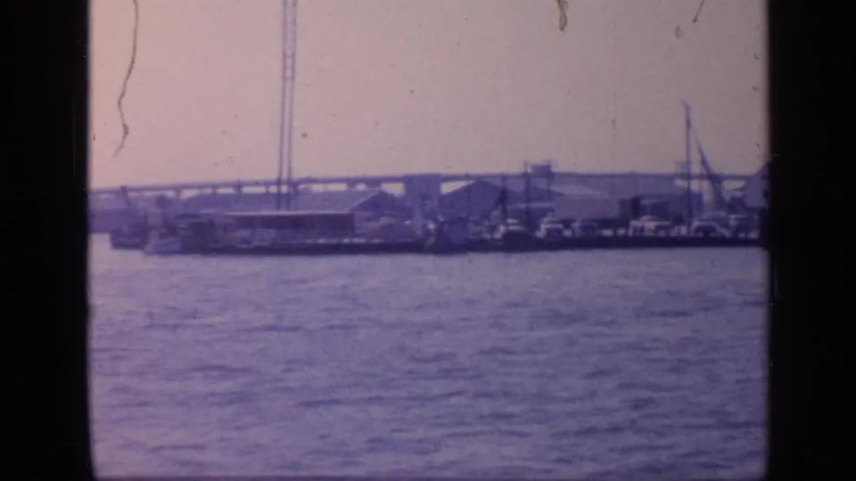 1968 Florida 8mm frame showing a distant seaport across rippled water, with boats, low waterfront buildings, cranes, and a long bridge on the horizon.