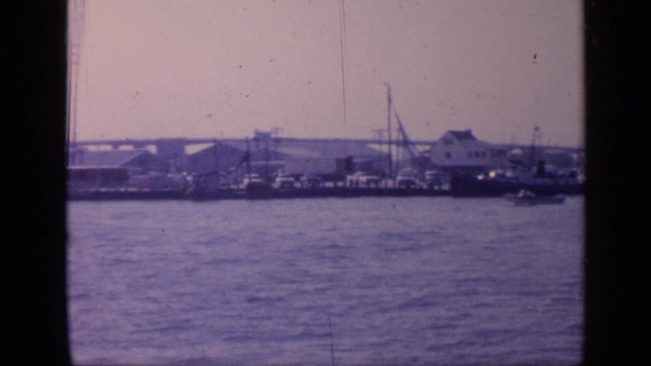 Wide 1968 Florida harbor view from the water, showing docked vessels, waterfront structures, crane-like equipment, and a long bridge in the distance.