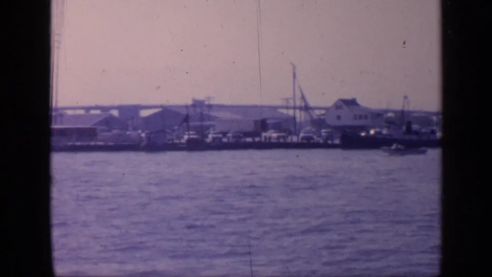 Wide 1968 Florida harbor view from the water, showing docked vessels, waterfront structures, crane-like equipment, and a long bridge in the distance.