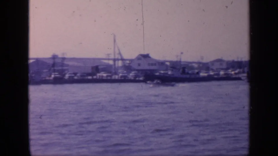 1968 Florida waterfront frame with a white dockside building, small boats, crane-like structures, and open water seen from a home-movie vantage point.