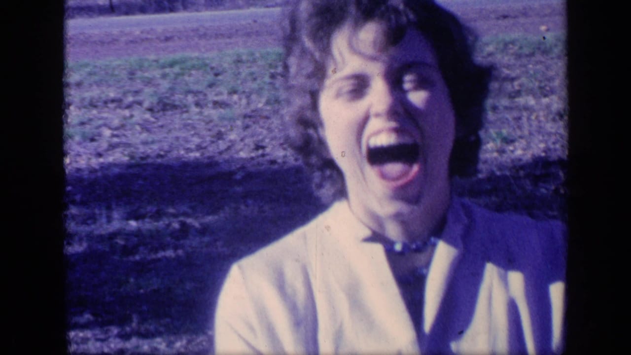 Young woman in a light jacket laughing outdoors near grass and trees.