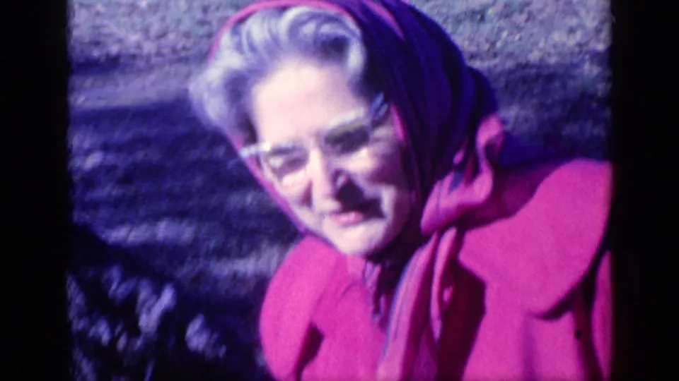 Older woman in glasses, a pink coat, and headscarf smiles toward the camera in 1968 Florida 8mm footage.