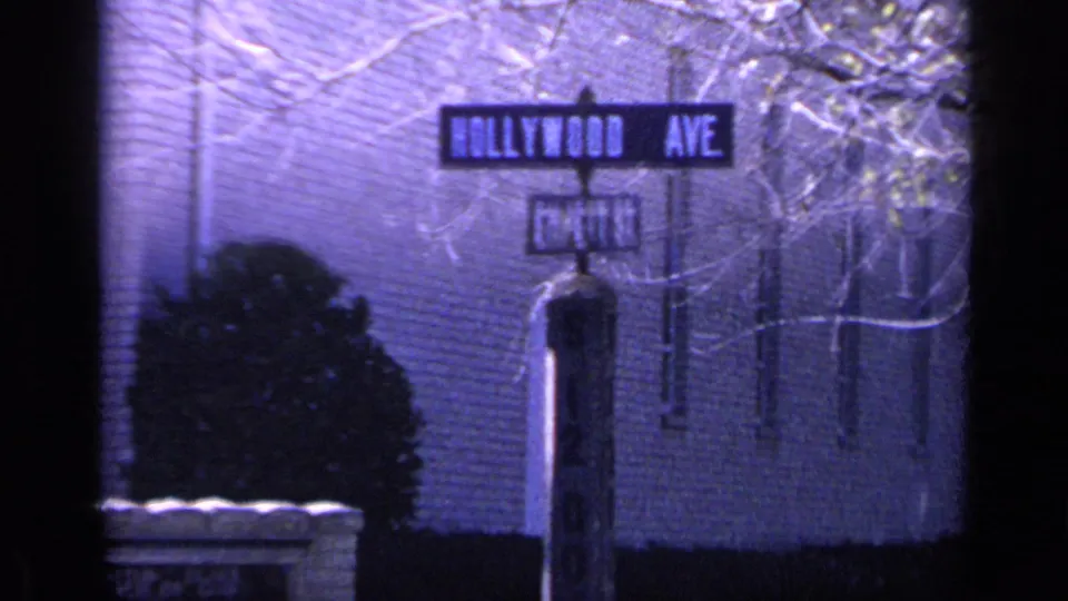 1968 Florida 8mm frame showing a Hollywood Ave street sign with a cross-street sign, brick wall, shrub, and bare branches.